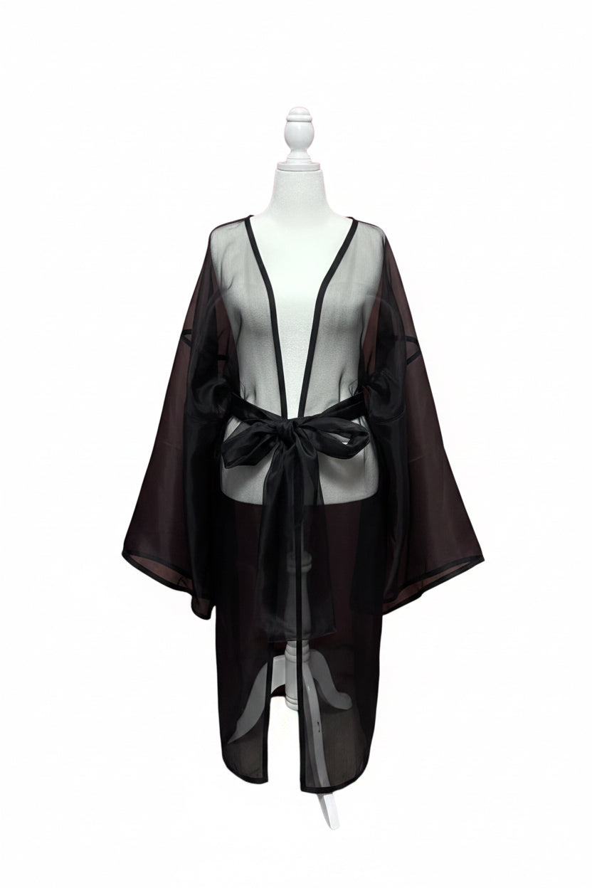 A black sheer mesh robe or kimono-style cover-up displayed on a white mannequin torso, with a deep V-neck and self-tie belt, set against a red curtain backdrop.